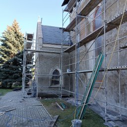 IS-International service - Budynek z szarej elewacją w trakcie remontu, otoczony rusztowaniami, widoczne deski, wiadro i materiały budowlane na trawie, częściowo zakryty czarną membraną dachową.