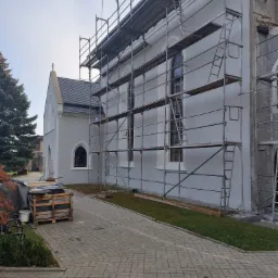 Kościół w trakcie remontu elewacji, widoczne rusztowania, paleta z materiałami budowlanymi i fragment chodnika z kostki brukowej.