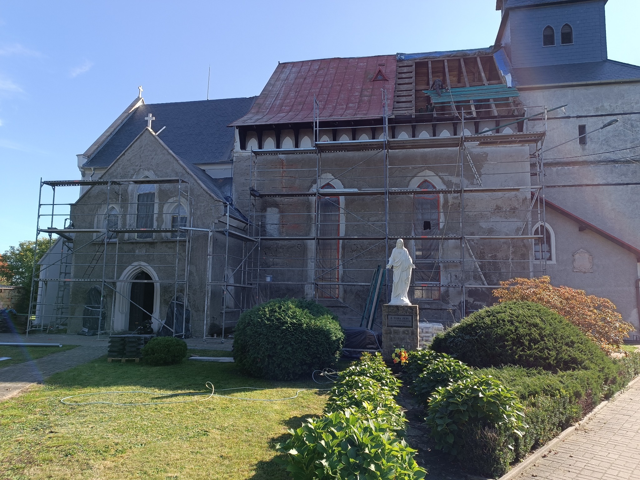 Kościół w trakcie remontu dachu, widoczne rusztowania, odsłonięta konstrukcja dachu i częściowo położone nowe pokrycie, z przodu pomnik.