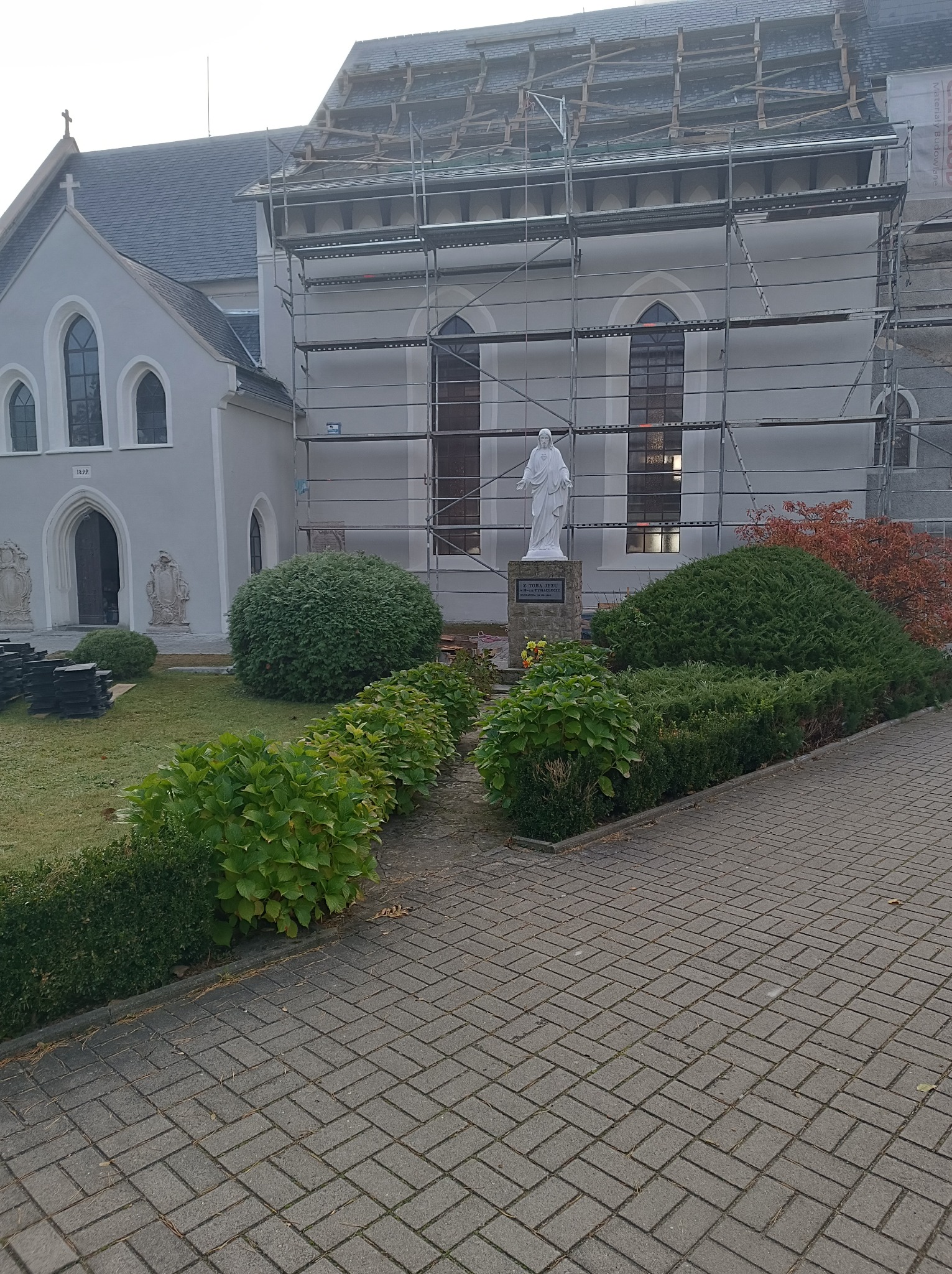 Kościół w trakcie remontu dachu, widoczne rusztowania, część dachu już pokryta nowym materiałem, na pierwszym planie rzeźba na tle zieleni.