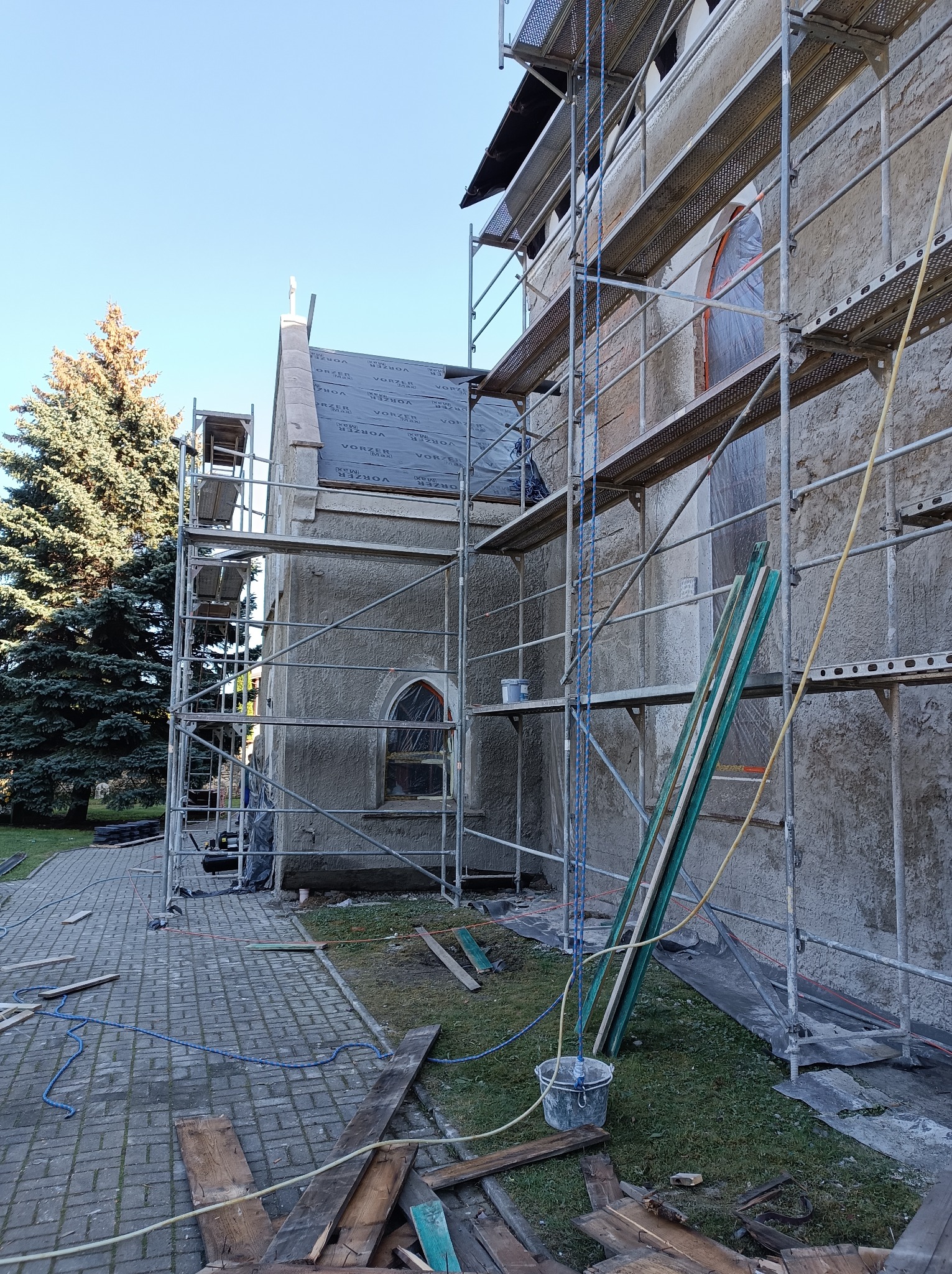 Budynek z szarej elewacją w trakcie remontu, otoczony rusztowaniami, widoczne deski, wiadro i materiały budowlane na trawie, częściowo zakryty czarną membraną dachową.