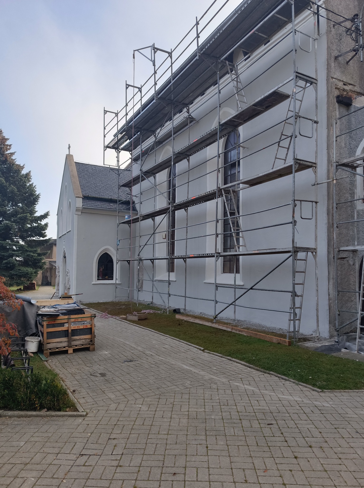 Kościół w trakcie remontu elewacji, widoczne rusztowania, paleta z materiałami budowlanymi i fragment chodnika z kostki brukowej.