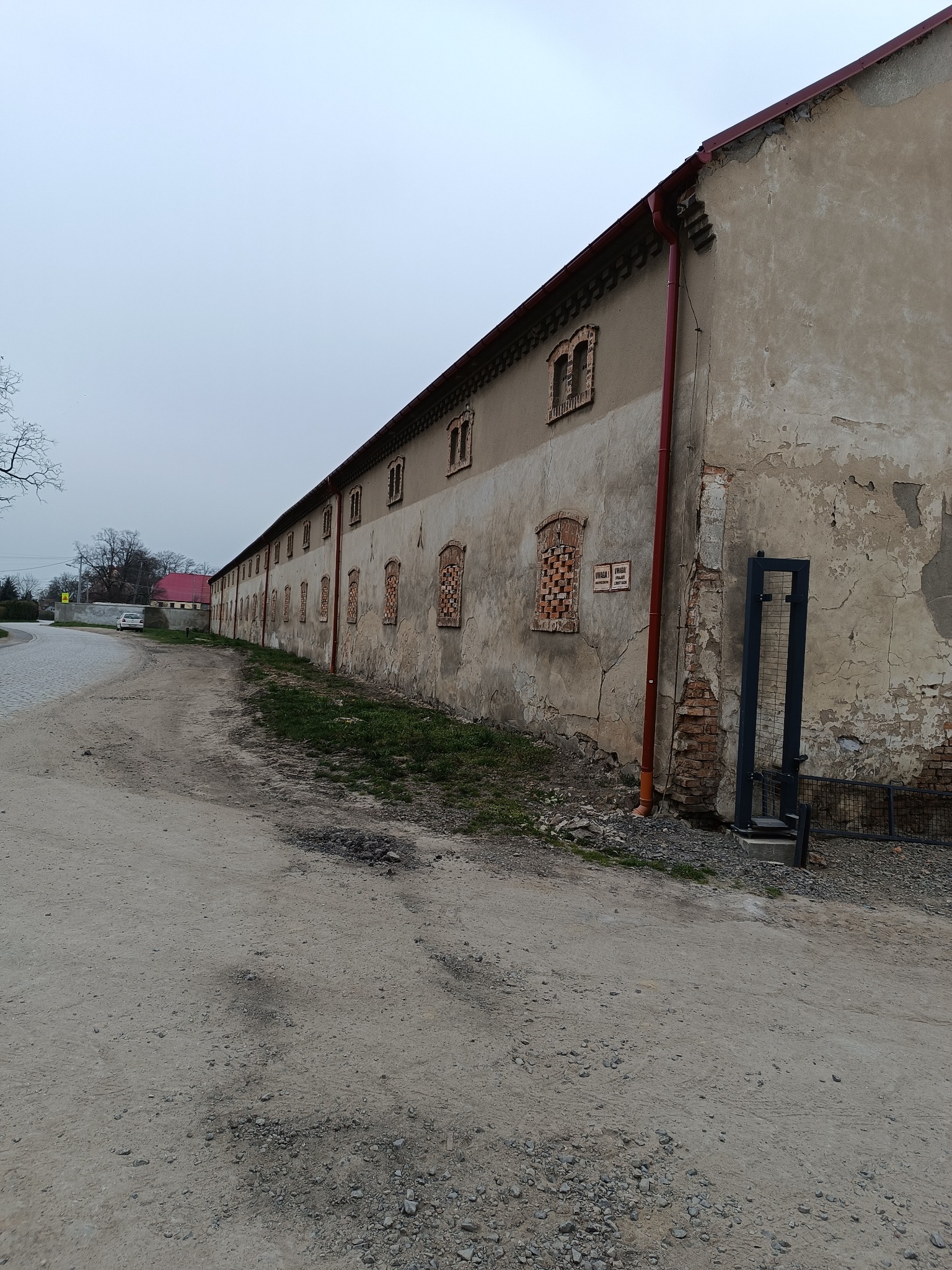 Długi budynek gospodarczy z ceglanymi detalami okiennymi i rynnami, widoczny z boku na tle pochmurnego nieba i żwirowej drogi.