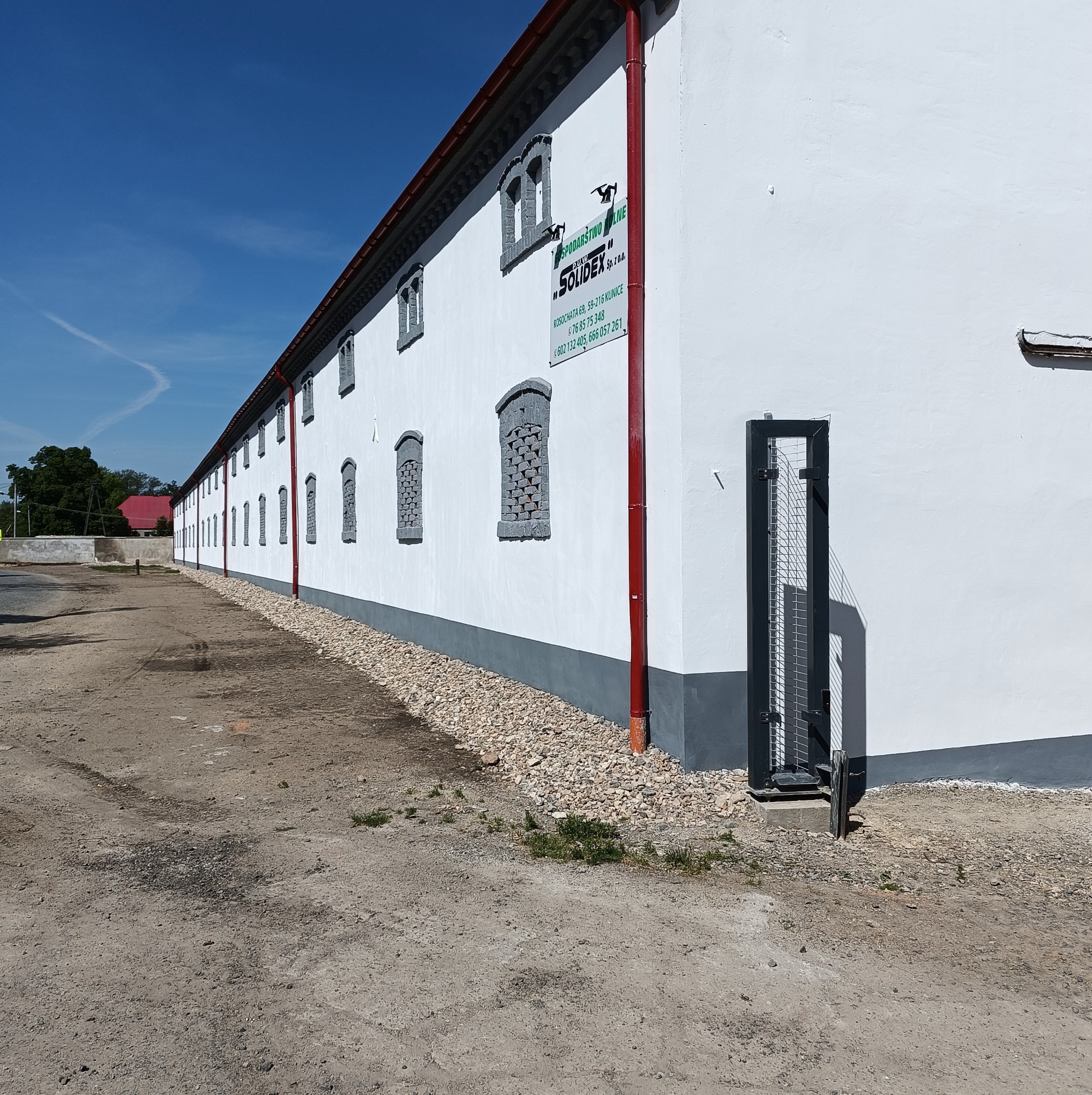 Długi, biały budynek gospodarczy z szarymi detalami okien i czerwoną rynną, z logo firmy Solidex na ścianie, widziany z perspektywy ziemi.