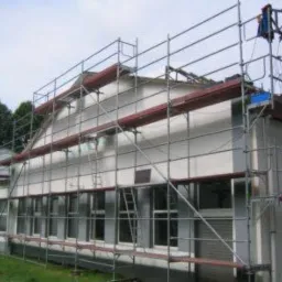 Budynek w trakcie remontu, otoczony rusztowaniami, z widocznymi elementami dachu w trakcie naprawy. Na górze rusztowania widać podnośnik z niebieskim pojemnikiem.