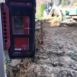 Elektroniczny niwelator zamontowany na łacie, w tle wykop pod fundamenty i pracująca koparka.