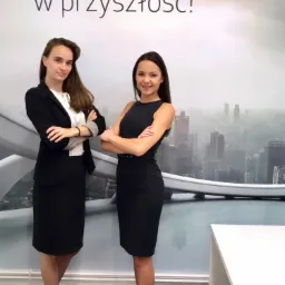 Dwie kobiety w eleganckich strojach biznesowych stojące na tle ściany z napisem 'Zespół Ekspercki Open Finance. Lepsze spojrzenie w przyszłość!' i panoramą miasta.