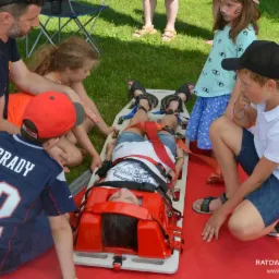 Grupa dzieci i dorosłych ćwiczy zabezpieczanie poszkodowanego na desce ortopedycznej na trawie, część osób siedzi wokół, jedna osoba leży na desce z założonym kołnierzem ortopedycznym i pasami.