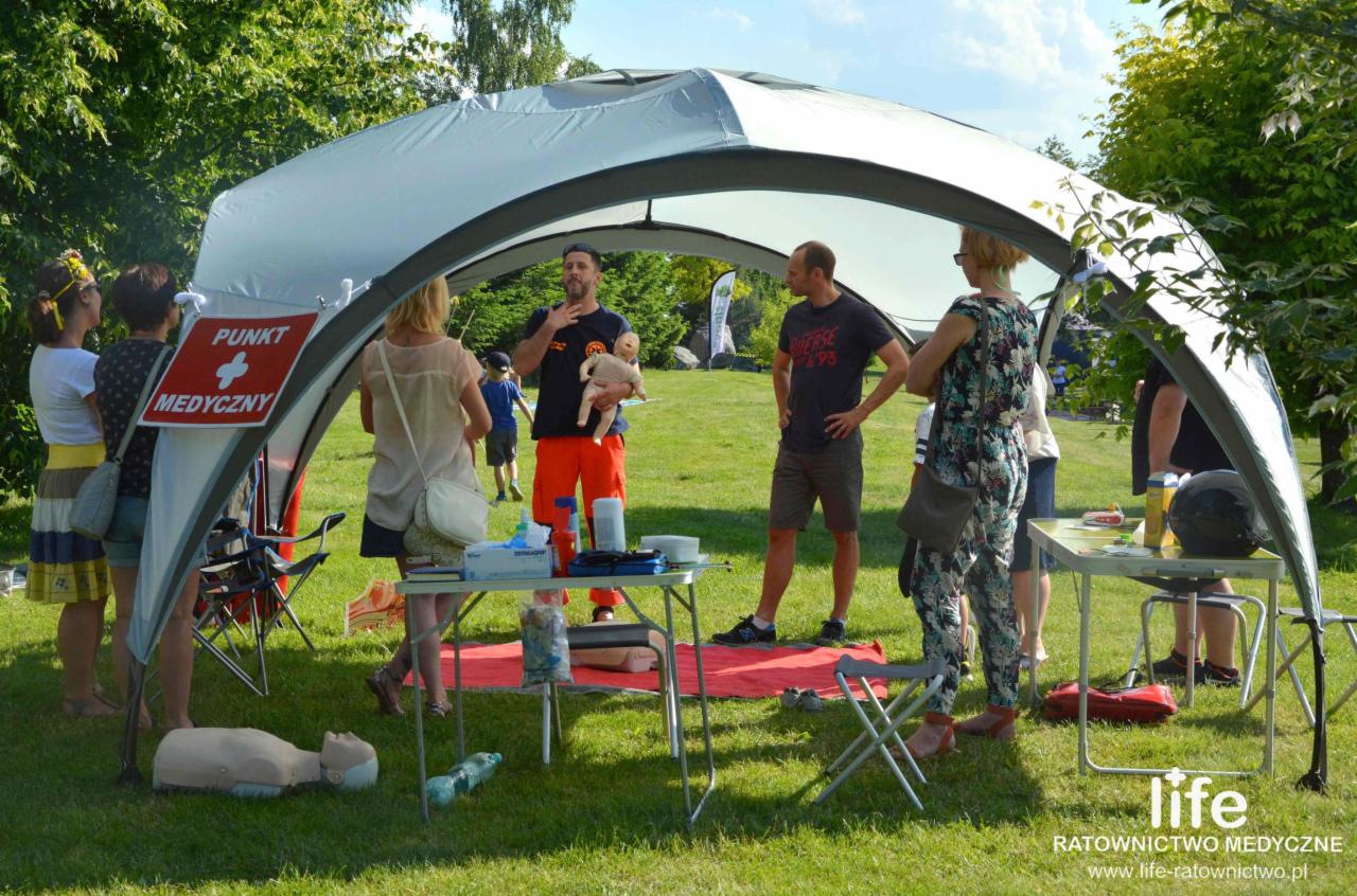 Instruktor w pomarańczowych spodniach demonstruje na manekinie niemowlęcia techniki ratownicze pod namiotem oznaczonym jako 'Punkt Medyczny', otoczony grupą słuchaczy na zielonym trawniku.