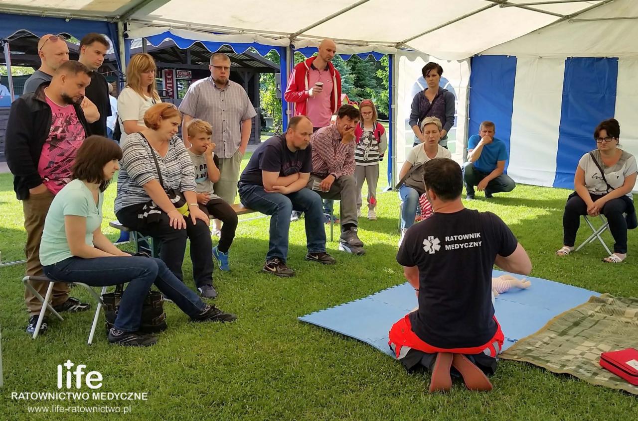 Instruktor medyczny demonstruje resuscytację krążeniowo-oddechową na manekinie przed grupą osób siedzących i stojących pod namiotem.