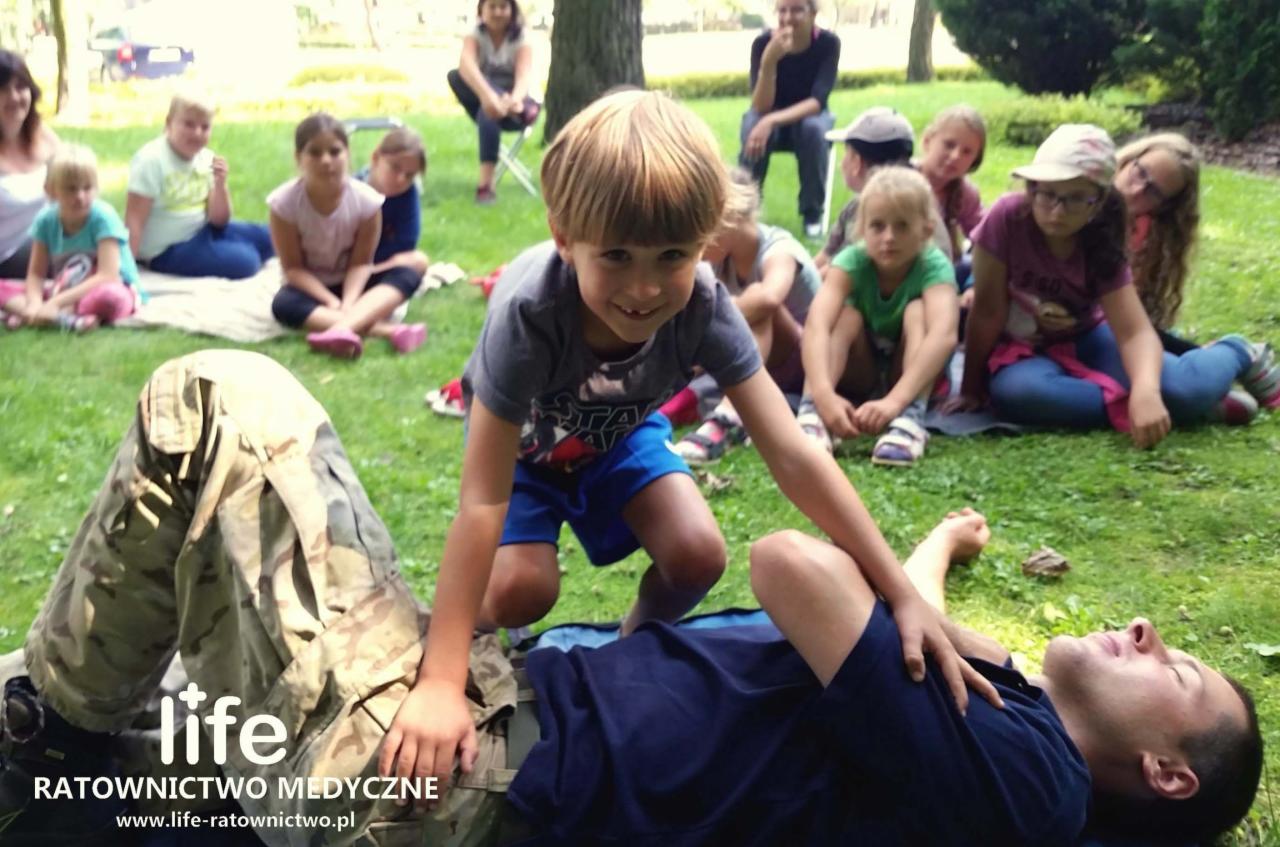Chłopiec uczy się resuscytacji na manekinie, wokół siedzą dzieci na trawie, w tle drzewa i budynki.
