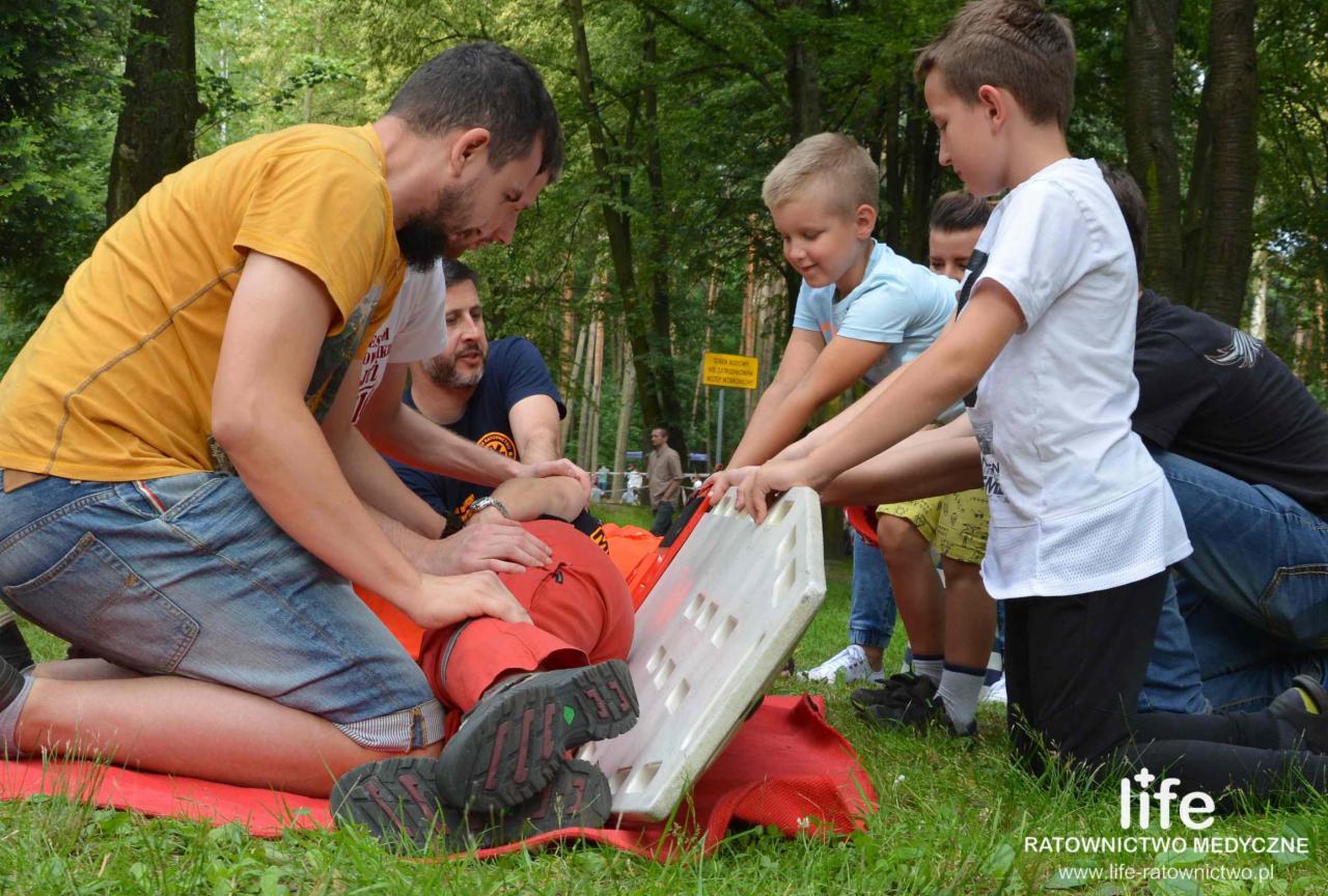Grupa osób, w tym dzieci, ćwiczy przenoszenie osoby poszkodowanej na desce ortopedycznej podczas zajęć na trawie w plenerze.