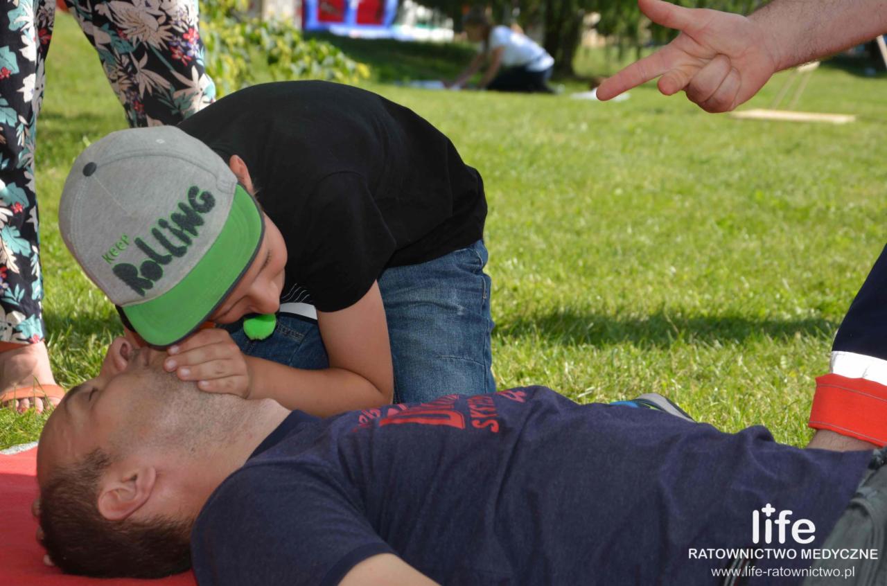 Chłopiec w czarnej koszulce i czapce Rolling udziela pierwszej pomocy, używając maski resuscytacyjnej na leżącym mężczyźnie na trawie, z widoczną dłonią wskazującą z góry i logiem 'life ratownictwo...