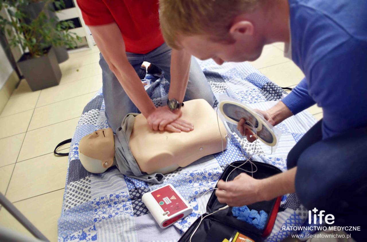 Man performing chest compressions on a CPR training dummy while another man prepares the AED pads, placed on a blue and white patterned blanket.