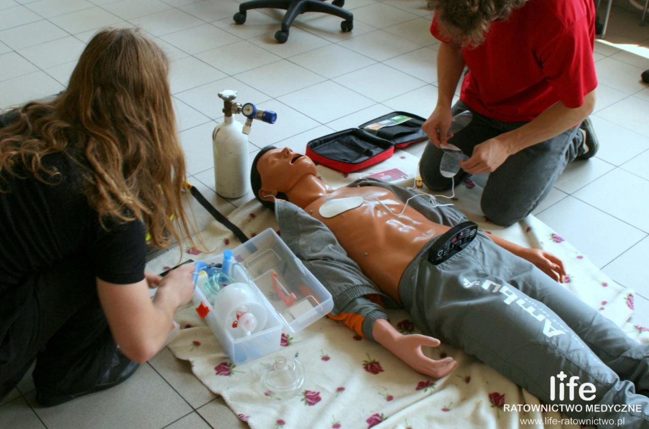 Instruktorzy demonstrują procedury pierwszej pomocy na manekinie z podłączonym sprzętem medycznym, w tym butlą z tlenem i defibrylatorem treningowym, podczas zajęć.