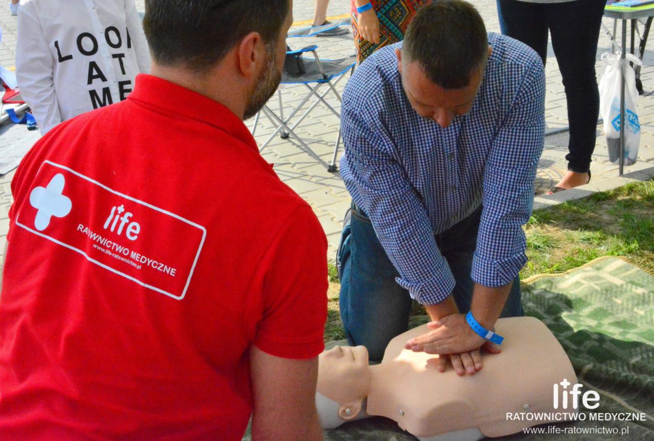 Instruktor w czerwonej koszulce z logo ratownictwa medycznego obserwuje mężczyznę w niebieskiej koszuli, który wykonuje resuscytację krążeniowo-oddechową na manekinie podczas szkolenia na zewnątrz.