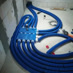 Niebieskie, karbowane przewody wentylacyjne rozprowadzone w surowym pomieszczeniu z bloczków, podłączone do niebieskiej skrzynki rozdzielczej, widoczne narzędzia i akcesoria montażowe.