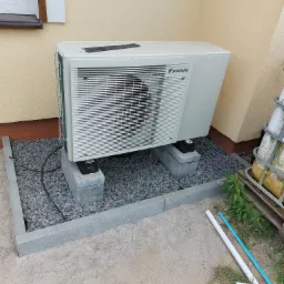 Zewnętrzna jednostka pompy ciepła Daikin, zamontowana na betonowych blokach, otoczona szarym żwirem i obrzeżem, z widocznymi rurami i elementami instalacji w tle.