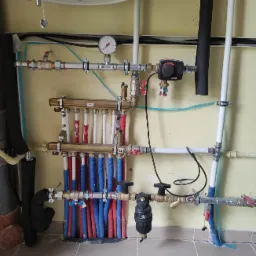 Rozdzielacz ogrzewania podłogowego z licznymi rurami w kolorach czerwonym i niebieskim, zamontowany na ścianie, z widocznym manometrem i filtrem.