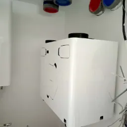 Biała centrala wentylacyjna z rekuperacją zamontowana na ścianie, widoczne rury wentylacyjne z kolorowymi zakończeniami oraz przewody instalacyjne.