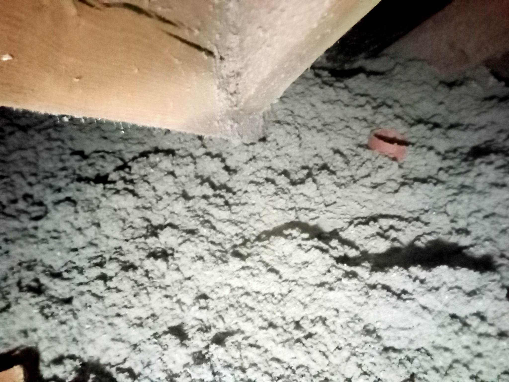 Szara, włóknista izolacja poddasza, widoczna spod drewnianej belki stropowej, z fragmentem pomarańczowej rurki. Nierównomierna powierzchnia izolacji z widocznymi cieniami.