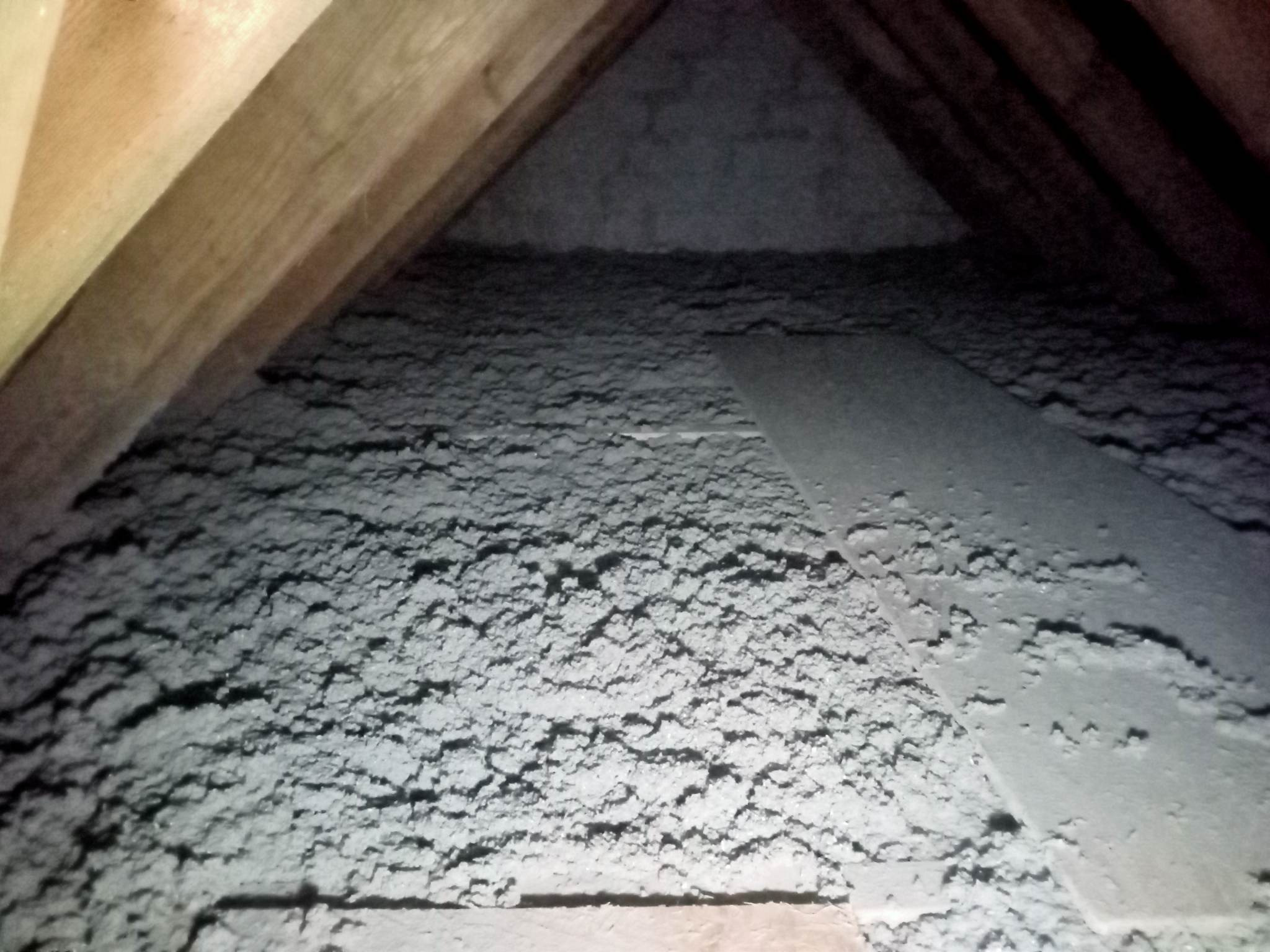 Szara warstwa izolacji z celulozy wypełniająca przestrzeń poddasza między drewnianymi krokwiami, widoczne nierówności powierzchni.