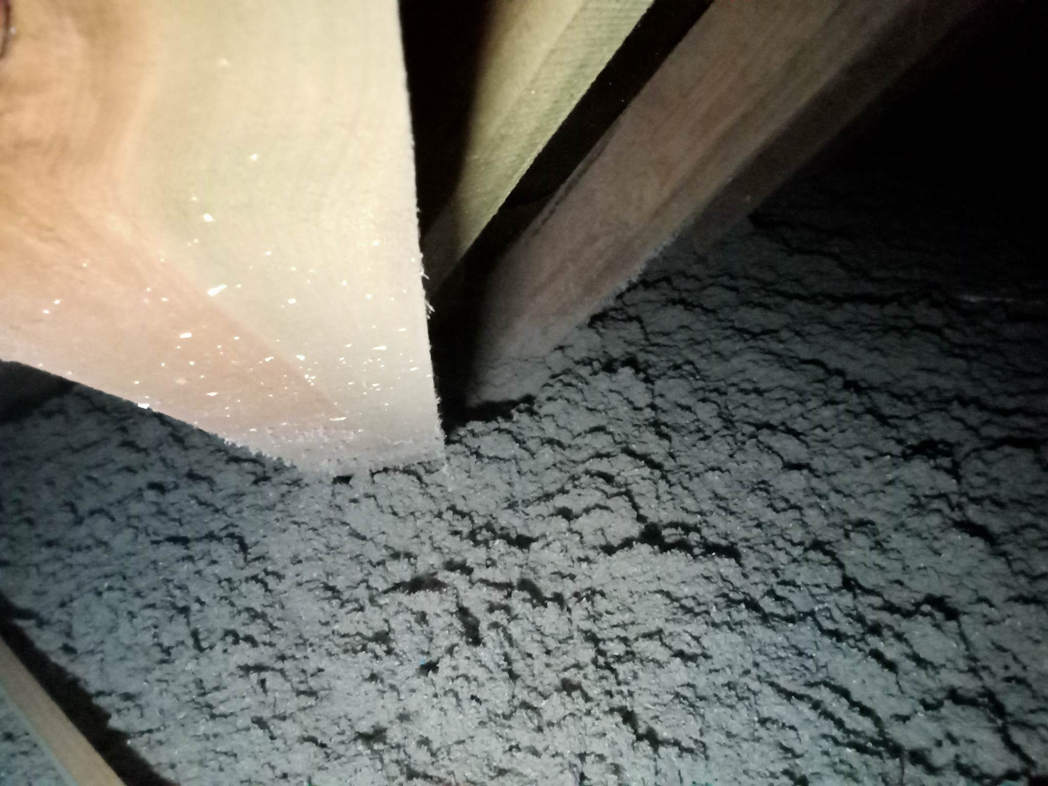 Szara wełna mineralna wypełniająca przestrzeń między drewnianymi belkami stropu poddasza, widok z dołu w słabym oświetleniu.