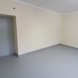 Puste pomieszczenie z szarą podłogą wylewką, kremowe ściany, szara futryna drzwiowa z czarną klamką i biały sufit.