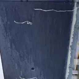 Ciemna elewacja z wąskich, prostokątnych płytek, widoczne kable i otwór, prace w toku.