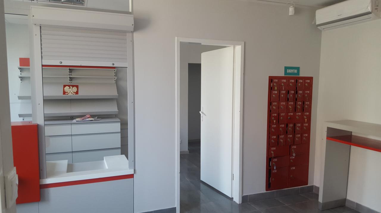 Wnętrze placówki pocztowej po remoncie: widok na stanowisko obsługi klienta z roletą i regałami, otwarte drzwi do zaplecza oraz czerwone skrzynki pocztowe z numerami na ścianie.