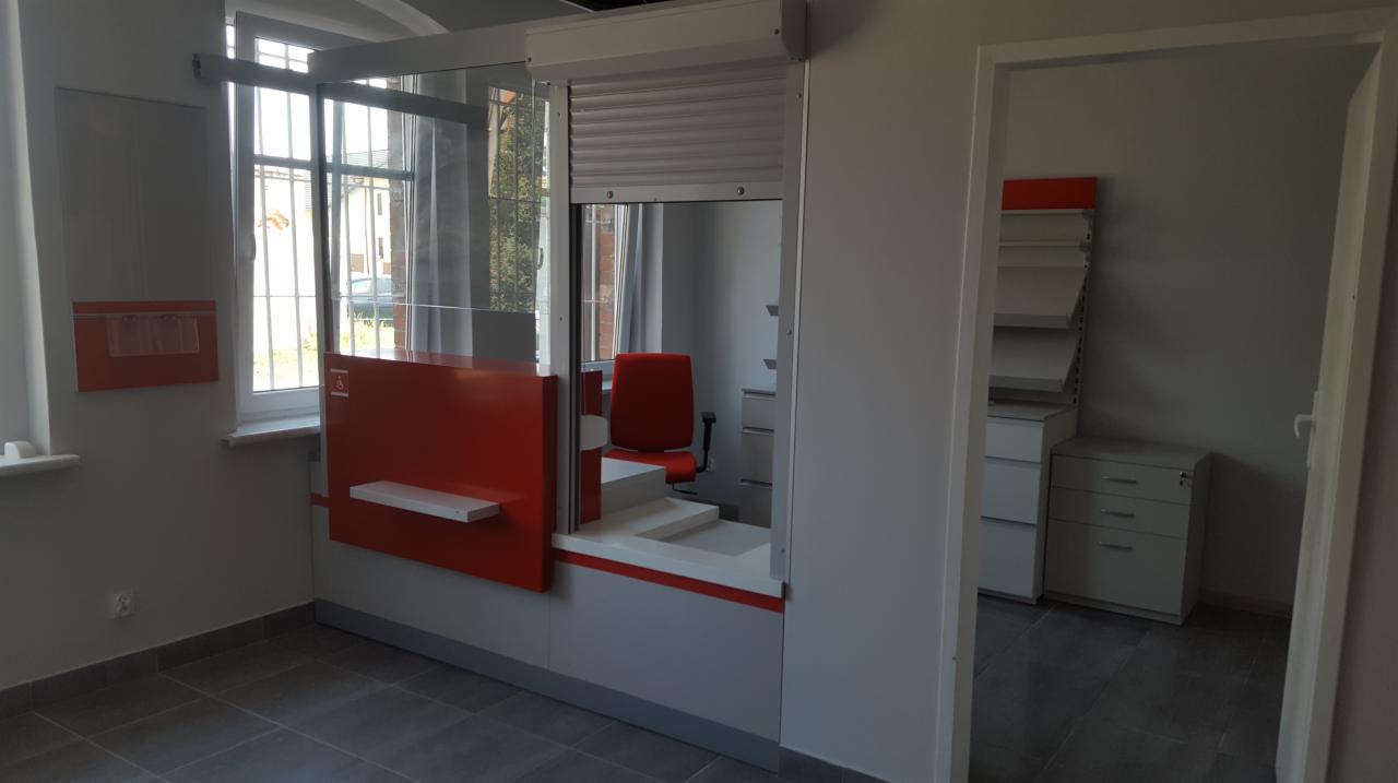 Nowoczesne wnętrze punktu obsługi klienta z pomarańczowymi akcentami, szarą roletą i oknem z kratami, widok na biuro z regałami w tle.