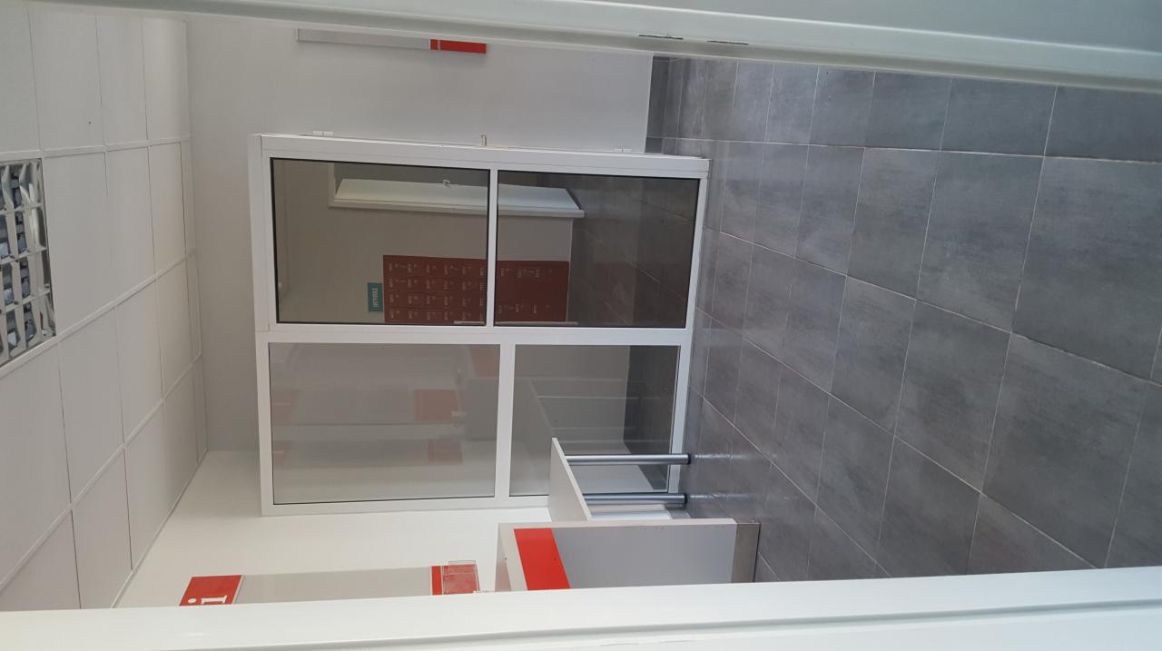 Wykończone wnętrze korytarza z szarą, kwadratową glazurą na podłodze, białą, przeszkloną ścianką działową z aluminium i białymi panelami ściennymi.