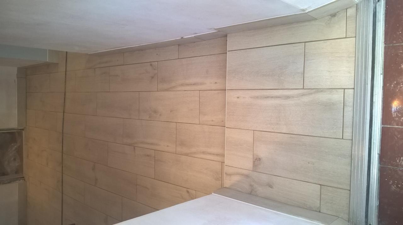 Ułożone poziomo płytki imitujące drewno na ścianie, widoczny fragment sufitu i narożnik obłożony płytkami, wykończony srebrną listwą.