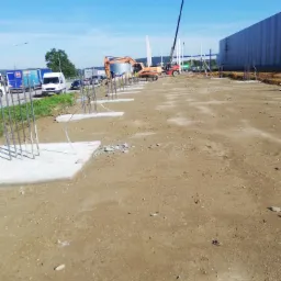 Plac budowy z przygotowanymi fundamentami pod budynek, widoczny dźwig, koparka gąsienicowa, wstępnie zbrojone słupy i zaparkowane busy w tle, słoneczny dzień.