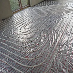 Rury ogrzewania podłogowego ułożone na folii z siatką w nowym budynku, widoczne okno i surowy beton na ścianach.