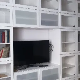 Biała zabudowa meblowa z licznymi szafkami i półkami, niektóre z książkami, z wbudowanym telewizorem, widok na fragment pokoju przez otwarte drzwi.