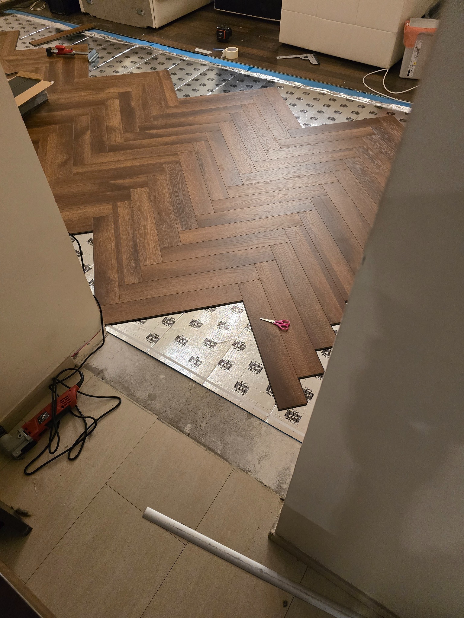 Układanie paneli w jodełkę na podkładzie izolacyjnym. Widoczne narzędzia: nożyczki i szlifierka kątowa. Częściowo ułożona podłoga w salonie.