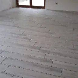 Ułożona podłoga z płytek imitujących drewno w jasnym odcieniu szarości, z widocznymi krzyżykami dystansowymi, w surowym pomieszczeniu z dużym oknem balkonowym.