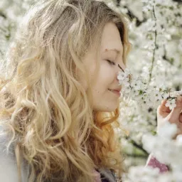 Blondwłosa kobieta z zamkniętymi oczami wącha kwitnącą gałązkę drzewa owocowego, otoczona białymi kwiatami, delikatne oświetlenie.