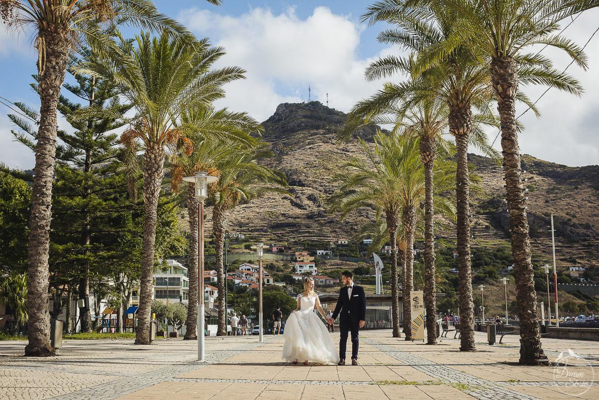 Para młoda trzymająca się za ręce spaceruje po placu z palmami, w tle góra z zabudowaniami.