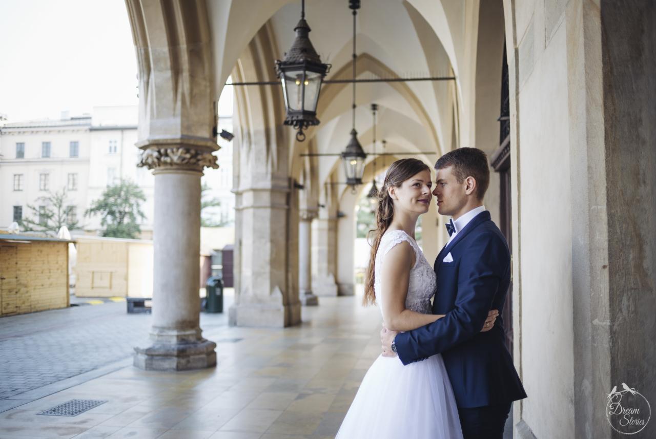 Para młoda w objęciach, na tle arkad Sukiennic w Krakowie, panna młoda w koronkowej sukni i pan młody w granatowym garniturze z muszką.