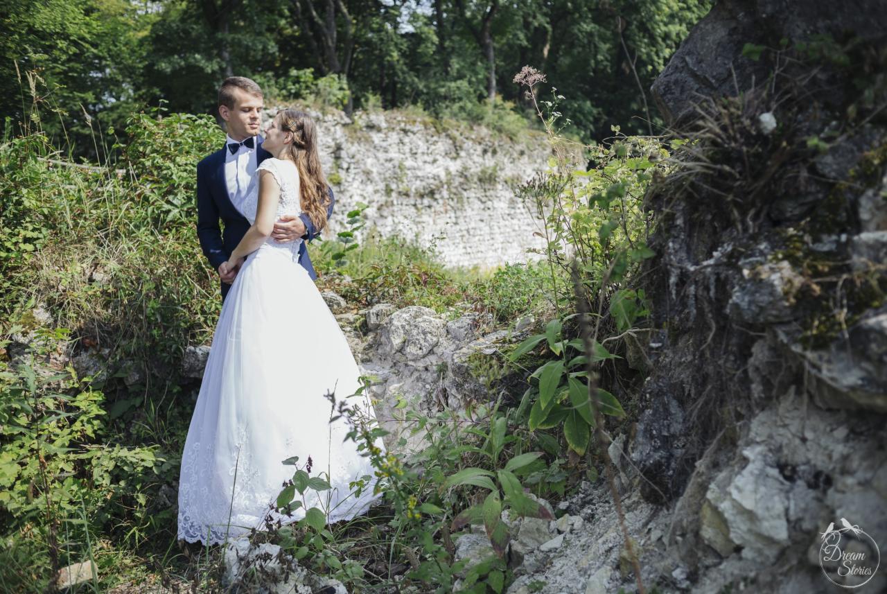Para młoda w objęciach na tle kamiennego muru i zieleni; panna młoda w białej sukni z koronkową górą, pan młody w granatowym garniturze z muchą.