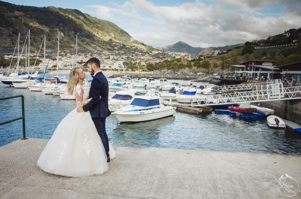 Para młoda w objęciach na tle portu jachtowego, góry i zabudowań miejskich w oddali. Panna młoda w białej sukni z koronkowymi aplikacjami, pan młody w granatowym garniturze.