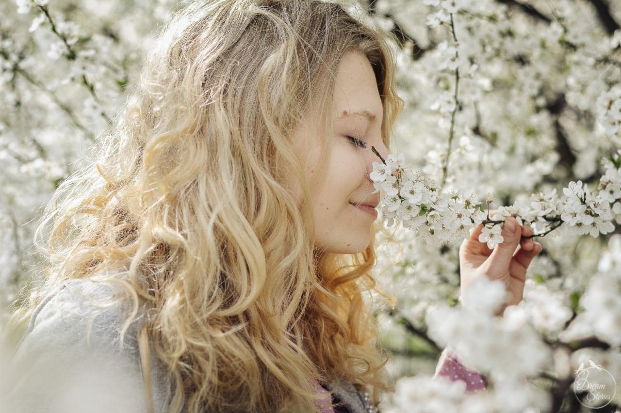 Blondwłosa kobieta z zamkniętymi oczami wącha kwitnącą gałązkę drzewa owocowego, otoczona białymi kwiatami, delikatne oświetlenie.