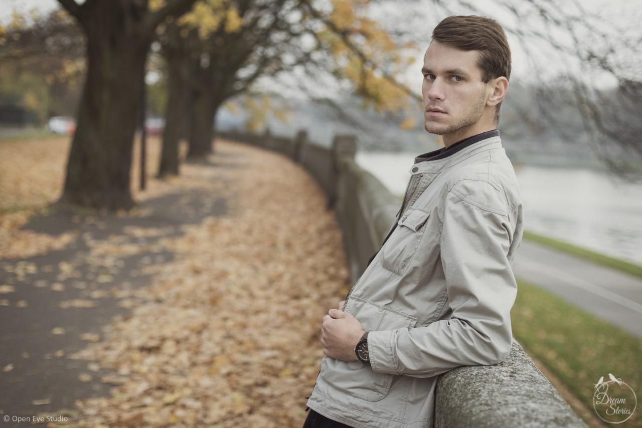 Młody mężczyzna w jasnej kurtce opiera się o kamienne ogrodzenie nad rzeką, jesienne drzewa w tle, spogląda w bok.