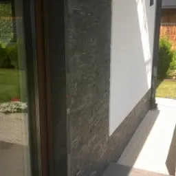 Fragment elewacji domu z ciemnoszarym kamieniem dekoracyjnym ułożonym w pasy, oświetlenie zewnętrzne w formie lampy na ścianie, widoczny fragment okna i trawnika.