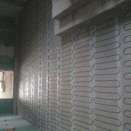 Instalacje elektryczne Łukasz Piekut - Ściana w trakcie montażu ogrzewania podłogowego ściennego, widoczne maty grzewcze z niebieskim przewodem układane pionowo na ścianie w surowym stanie, w tle okno.