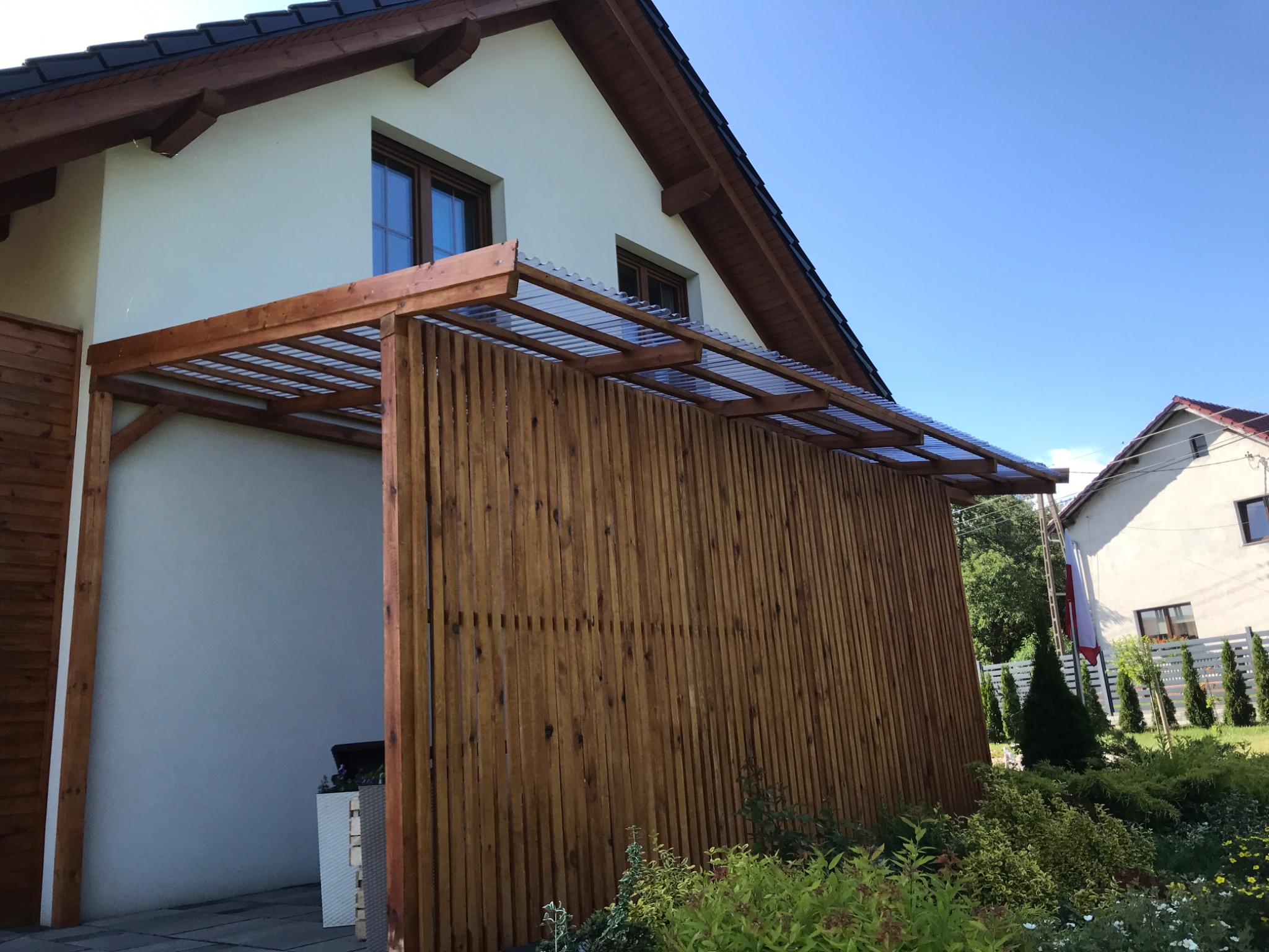 Drewniana konstrukcja z zadaszeniem z poliwęglanu przy domu w Niemodlinie, widok z zewnątrz na tle zadbanego ogrodu.