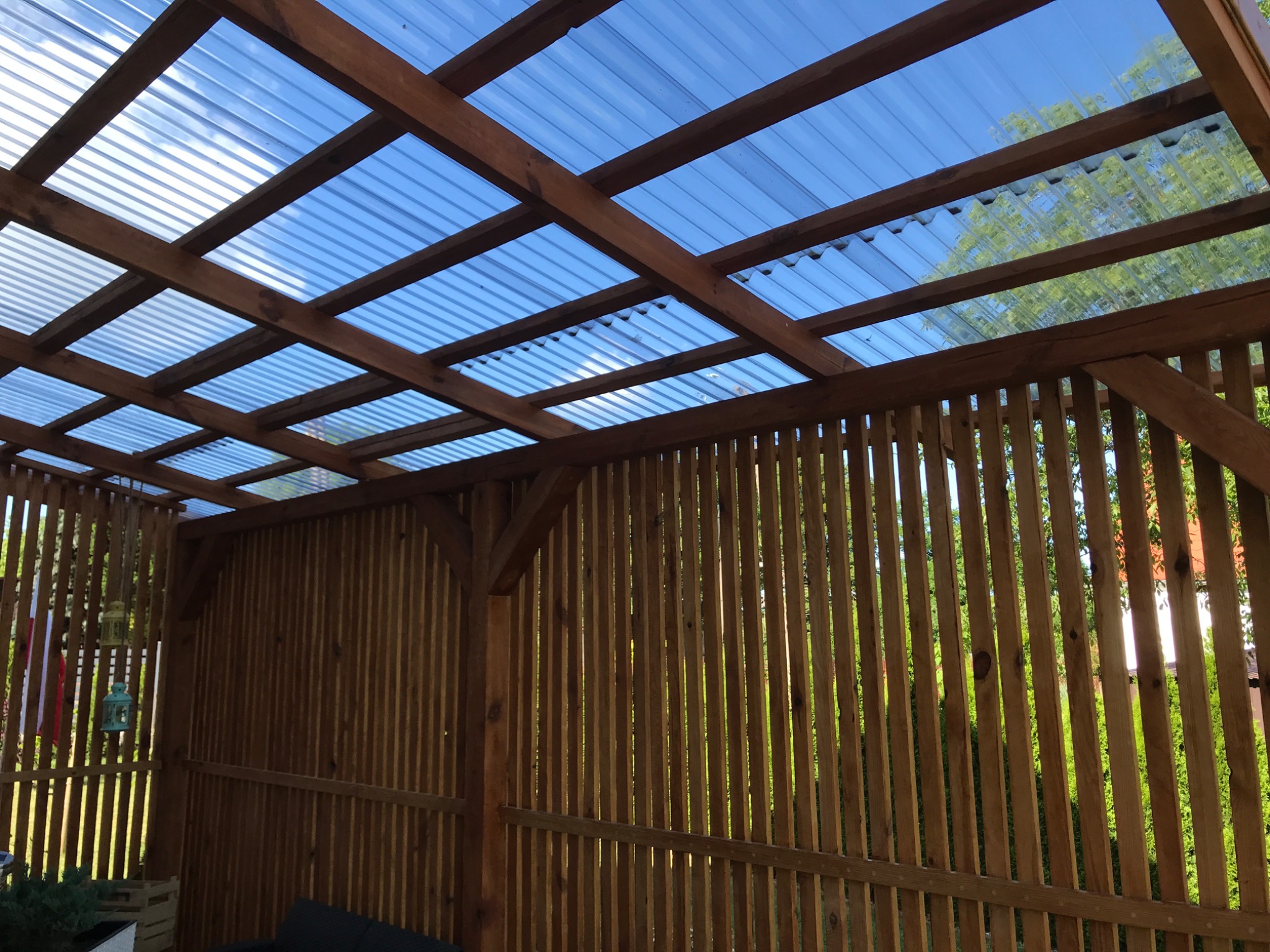 Drewniana konstrukcja z przezroczystym, falistym zadaszeniem, ukazująca błękitne niebo, boczne ścianki z pionowych listew, w tle zieleń drzew.