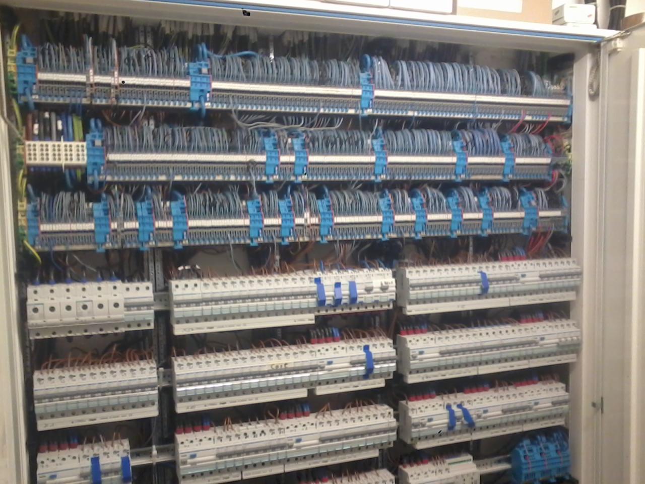 Otwarta szafa elektryczna z licznymi przewodami, listwami zaciskowymi i zabezpieczeniami, uporządkowanymi w rzędach, widoczne niebieskie elementy mocujące.